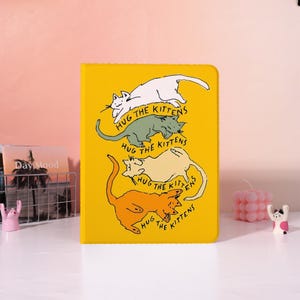 May include: A bright yellow notebook featuring cartoon kittens in white, green, beige, and orange. The text "HUG THE KITTENS" is repeated below each kitten. The notebook is on a white surface with decorative items in the background.