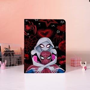 May include: A rectangular tablet case featuring a black background with a graphic illustration of two masked figures in white, red, and blue suits. The design includes red spider webs, stars, a heart, and spider motifs scattered across the surface.