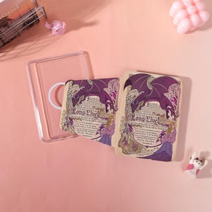 May include: A clear phone case with a printed insert. The insert features a purple dragon, a woman in a purple dress, and the text "Long Live!". The design is set against a parchment-like background with decorative elements. The case is shown on a pink surface.