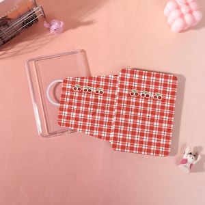 May include: A clear plastic phone case and two red and white checkered cardholders with cherry designs. The items are on a pink surface, with small decorative items in the background.