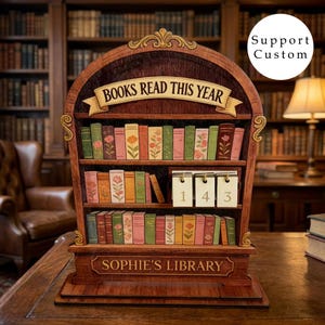 Könnte beinhalten: Ein hölzernes Bücherregal-Display mit der Aufschrift "BOOKS READ THIS YEAR" oben und "SOPHIE'S LIBRARY" an der Basis. Das Objekt enthält drei Regalebenen mit Miniatur-Buchrücken, die mit Blumenmustern verziert sind, sowie ein Zählwerk.