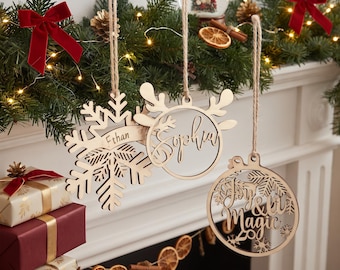 May include: Three laser-cut wooden Christmas ornaments hanging from twine. One is a snowflake with the name Ethan, one is a circular reindeer design with the name Sophia, and one is a circular ornament with the text Joy & Magic and snowflakes.