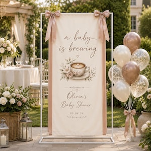 May include: A vertical cream-colored fabric banner on a white metal stand, featuring the text 'a baby is brewing', a coffee cup illustration, and 'welcome to Olivia's Baby Shower 09.08.26'. The banner is decorated with two dusty pink satin bows.