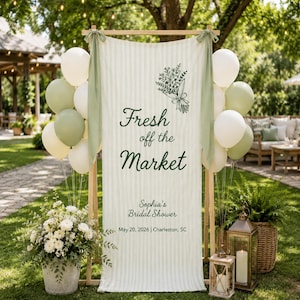 May include: A decorative sign with the text "Fresh off the Market" and "Sophia's Bridal Shower" is displayed outdoors. The sign is decorated with a floral illustration and green and white balloons. The date and location are also included.