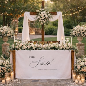 May include: A wooden wedding altar with a cross, draped with white fabric and adorned with white flowers. A table is decorated with flowers, candles, and the names "The Smith" and the wedding date.