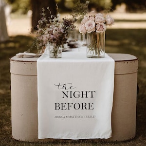 May include: A wedding table setting with a white linen runner featuring the text "the NIGHT BEFORE" in black script. The table is decorated with floral arrangements in glass vases. The table is set on two beige vintage hat boxes.