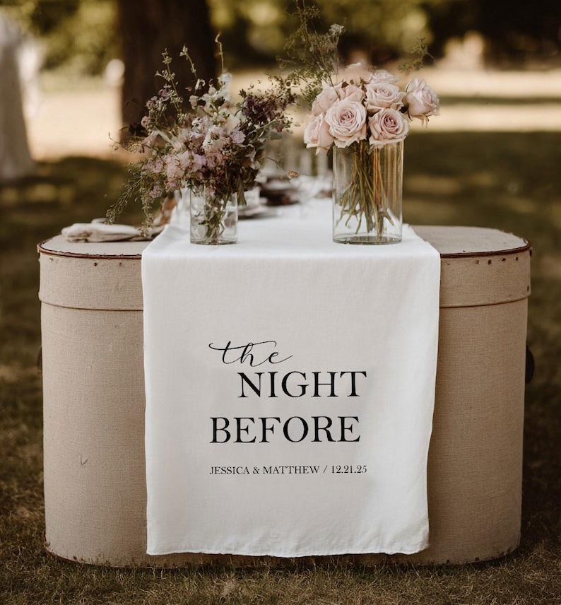 May include: A wedding table setting with a white linen runner featuring the text "the NIGHT BEFORE" in black script. The table is decorated with floral arrangements in glass vases. The table is set on two beige vintage hat boxes.
