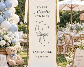 May include: A vertical cream-colored fabric banner suspended on a gold metal stand, featuring the black text TO THE MOON AND BACK, a crescent moon illustration with a bow, the name BABY CARTER, and the date 04.26.2026, accented by cream ribbons.