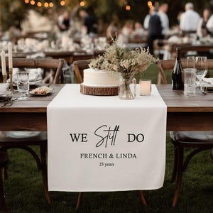 Könnte beinhalten: Ein weißer Tischläufer mit dem Text "WE Still DO FRENCH & LINDA 25 years" liegt auf einem Holztisch, der für eine Feier gedeckt ist. Ein Kuchen, Blumen und Kerzen sind auf dem Tisch angeordnet, mit Gästen im Hintergrund.