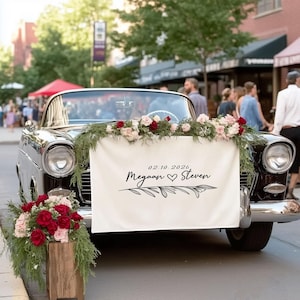 Pode incluir: Um carro conversível preto clássico decorado para um casamento. O carro tem uma faixa com o texto "02.10.2026, Meagan & Steven" e arranjos florais. Uma caixa de madeira com flores está em frente ao carro.