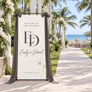 May include: A vertical cream-colored wedding welcome sign banner featuring the text "WELCOME TO OUR WEDDING", the initials "ED", the names "Emily & Daniel", and the date "SEPTEMBER 20, 2026", accented with a single orchid illustration and dark gray fabric bows.