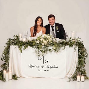 May include: A couple sits at a decorated table with a white tablecloth. The table is adorned with greenery, white flowers, candles, and glassware. The tablecloth features the initials "B" and "S" with the names "Brian & Sophia" and the date "Est. 2026".