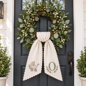 May include: A circular wreath made of green foliage and small white flowers, featuring a hanging beige fabric banner with scalloped edges. The banner displays an embroidered illustration of a baby carriage on one side and the letter O on the other.