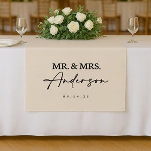 以下が含まれることがあります： 「MR. & MRS. Anderson」と日付「09.14.25」が黒で書かれたクリーム色のテーブルランナー。白いテーブルクロスの上に置かれ、背景にはフラワーアレンジメントとワイングラスがあります。