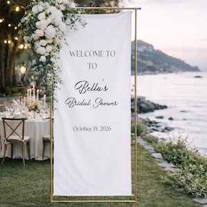 May include: A white banner on a gold frame, with the text "WELCOME TO TO Bella's Bridal Shower October 19, 2026." White flowers and greenery adorn the top. The background shows a table setting and a coastal view.