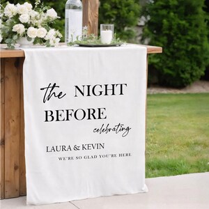 May include: A white fabric banner with black text that reads "the NIGHT BEFORE celebrating LAURA & KEVIN WE'RE SO GLAD YOU'RE HERE." The banner is draped over a wooden table, with floral arrangements and candles.