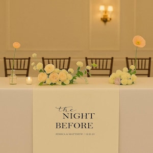 May include: A table setting with floral arrangements and a decorative table runner. The runner features the text "the NIGHT BEFORE" in elegant script, with names and a date below. Soft lighting and chairs are visible in the background.