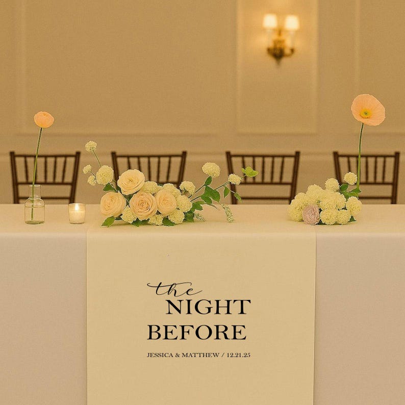 May include: A table setting with floral arrangements and a decorative table runner. The runner features the text "the NIGHT BEFORE" in elegant script, with names and a date below. Soft lighting and chairs are visible in the background.