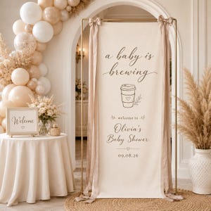 May include: A tall rectangular fabric banner on a gold metal stand featuring the text 'a baby is brewing', a coffee cup illustration, and 'welcome to Olivia's Baby Shower 09.08.26'. The banner is tied with beige ribbons at the top corners.