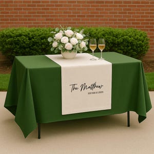 May include: A green tablecloth with a cream-colored table runner. The runner is printed with "The Matthew" and the date "EST 08.12.2025". A white floral arrangement and two wine glasses sit on the table. The table is set for a special event.