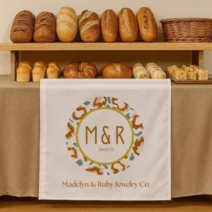 May include: A display of various baked goods, including loaves of bread, rolls, and pastries, arranged on wooden shelves and a table. A white table runner with the text "M&R Jewelry Co." and a circular logo is draped over the table.