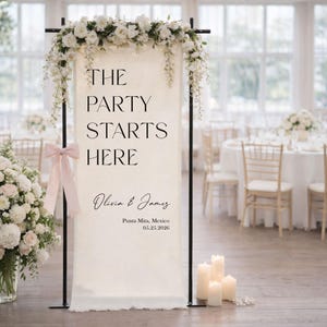 May include: A wedding reception sign with the text "THE PARTY STARTS HERE" in black font. The sign is decorated with white flowers and a pink bow. The names "Olivia & James" and the date 05.25.2026 are also displayed.
