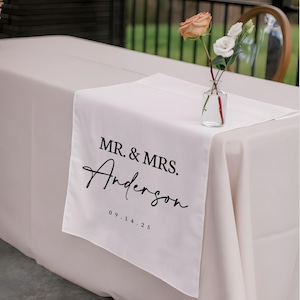 以下が含まれることがあります： 「MR. & MRS. Anderson」の文字と日付「09.14.25」が黒い筆記体で書かれた白いテーブルランナー。明るいベージュのテーブルクロスの上に置かれています。小さなガラスの花瓶に花が飾られています。