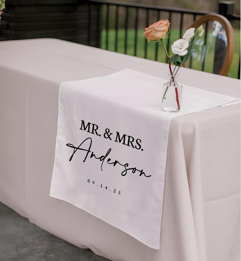 以下が含まれることがあります： 「MR. & MRS. Anderson」の文字と日付「09.14.25」が黒い筆記体で書かれた白いテーブルランナー。明るいベージュのテーブルクロスの上に置かれています。小さなガラスの花瓶に花が飾られています。