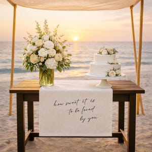 May include: A two-tiered white wedding cake with floral accents sits on a white cake stand. A table runner with the text "how sweet it is to be loved by you" is displayed on a dark wooden table. A floral arrangement and the ocean are in the background.