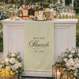 May include: A white bar with various bottles of liquor and wine, glassware, and a striped runner with the text "MR & MRS Munsch Est. 2026". The bar is decorated with flowers, lemons, and lanterns, creating a festive atmosphere.