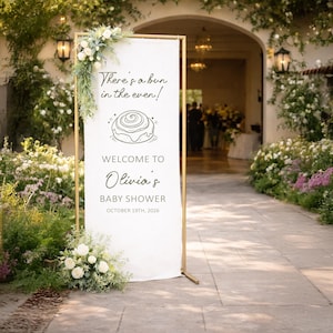 May include: A white sign with gold frame announces "Olivia's Baby Shower" with the text "There's a bun in the oven!" and a cinnamon roll illustration. The sign is decorated with white flowers and greenery, set outdoors.