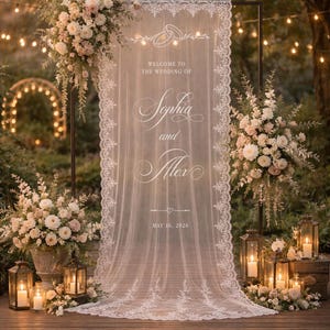 May include: A white lace wedding welcome sign hanging on a black metal frame. The fabric features a graphic of two interlocking rings, the text "WELCOME TO THE WEDDING OF Sophia and Alex", a small heart icon, and the date "MAY 16, 2026" printed in white.