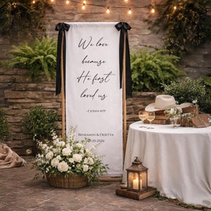 May include: A white wedding banner with black ribbon detailing features the text "We love because He first loved us" and the Bible verse 1 John 4:19. The banner is part of a wedding scene with floral arrangements and a table setting.