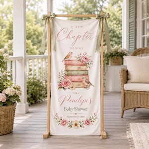 May include: A vertical fabric banner suspended from a wooden frame with sage green ribbons, featuring the text A NEW CHAPTER BEGINS, Penelope's Baby Shower, and September 14, 2026, decorated with a stack of books, birds, and pink floral illustrations.