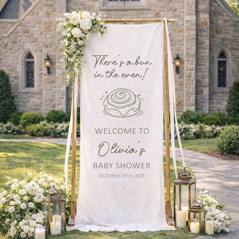 May include: A white fabric baby shower sign with the text "There's a bun in the oven!" and "Welcome to Olivia's Baby Shower October 19th, 2026." The sign is decorated with flowers and lanterns.