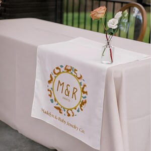 May include: A white table runner with a gold-colored circular logo featuring the letters "M&R" and the text "Madelyn & Ruby Jewelry Co." A small glass vase holds a rose and white flowers. The table is covered with a light beige tablecloth.