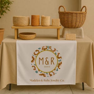 May include: A display of woven baskets and containers in various sizes and colors, arranged on a wooden shelf and table. The table runner features a logo with the text "M&R Jewelry Co." and "Madelyn & Ruby Jewelry Co."