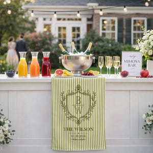 Può includere: Un bar per mimosa con bottiglie di champagne in un secchiello per il ghiaccio, vari succhi e un cartello con la scritta "Mimosa Bar". Un runner da tavolo a righe verdi e bianche mostra un monogramma e il nome "The Wilson".