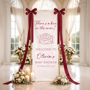 May include: A white baby shower welcome sign with burgundy text and accents. The sign reads "There's a bun in the oven!" and "Welcome to Olivia's Baby Shower October 19th, 2026." The sign is decorated with burgundy bows and ribbons.