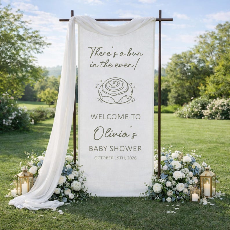 May include: A white banner with the text "There's a bun in the oven!" and "Welcome to Olivia's Baby Shower October 19th, 2026." The banner is part of a baby shower decoration, with floral arrangements and lanterns.