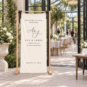 May include: A tall, rectangular, off-white fabric wedding welcome sign suspended from a light-colored wooden frame with black ribbons. The sign features black text reading: WELCOME TO OUR WEDDING, A&J, AVA & JAMES, joyfully invite you TO OUR WEDDING, AUGUST 24, 2026.