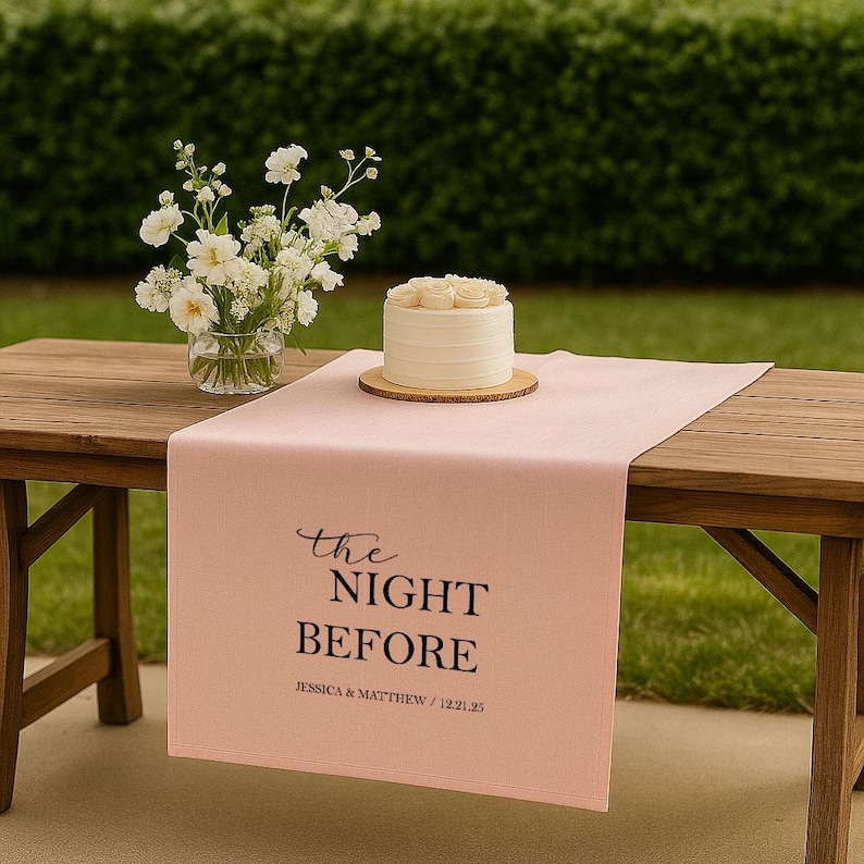 May include: A wooden table with a pink table runner inscribed with "The Night Before" and names and a date. A small white cake with rose decorations sits on a wooden base. A vase of white flowers is also on the table.