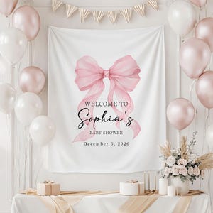 May include: A white backdrop with a large pink watercolor bow and the text "WELCOME TO Sophia's BABY SHOWER December 6, 2026." The scene is decorated with white and pink balloons, a table with gifts, and floral arrangements.