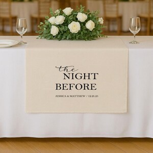 May include: A cream-coloured table runner with the text "the NIGHT BEFORE" in black script and block letters. Below the text is "JESSICA & MATTHEW / 12.21.25". The runner is on a white tablecloth, with a floral arrangement and wine glasses.