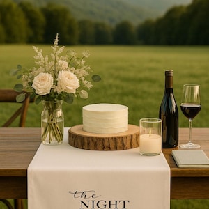May include: A wedding table setting featuring a white cake on a wooden base, a bottle of wine, a wine glass, and a candle. A vase of white roses and greenery is next to the cake. A white table runner reads "the NIGHT BEFORE".