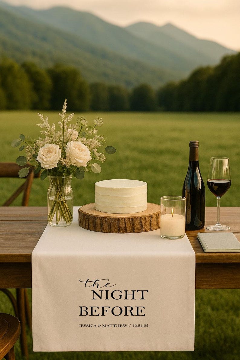 May include: A wedding table setting featuring a white cake on a wooden base, a bottle of wine, a wine glass, and a candle. A vase of white roses and greenery is next to the cake. A white table runner reads "the NIGHT BEFORE".