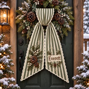 May include: A holiday wreath featuring green pine branches, pinecones, and white berries, adorned with a long, scalloped, sage green and white striped fabric ribbon. The ribbon displays the name Everett and small snowflake accents.