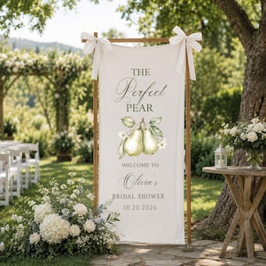 May include: A vertical white fabric banner suspended on a wooden frame with white ribbon bows at the top corners. The banner features a watercolor illustration of two pears and text reading THE PERFECT PEAR WELCOME TO Olivia's BRIDAL SHOWER 10.20.2026.