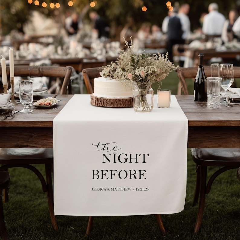 May include: A wedding reception table set with a white table runner bearing the words "the NIGHT BEFORE". The table is adorned with a cake, flowers, candles, and wine glasses. The date and names are also printed on the runner, creating a romantic atmosphere.