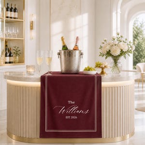 Puede incluir: Un camino de mesa burdeos con borde dorado y el texto "The Williams EST. 2026" se muestra en una barra. Una cubitera plateada contiene dos botellas. Copas de champán, flores y uvas también están en la barra.